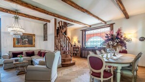 Un salón espacioso con una escalera de madera, un comedor con una mesa preparada para cuatro personas y un gran ventanal con detalles de hierro decorativo.