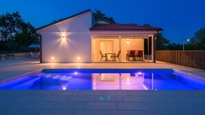 Una casa moderna con piscina, un patio cubierto con mobiliario exterior y una entrada bien iluminada.