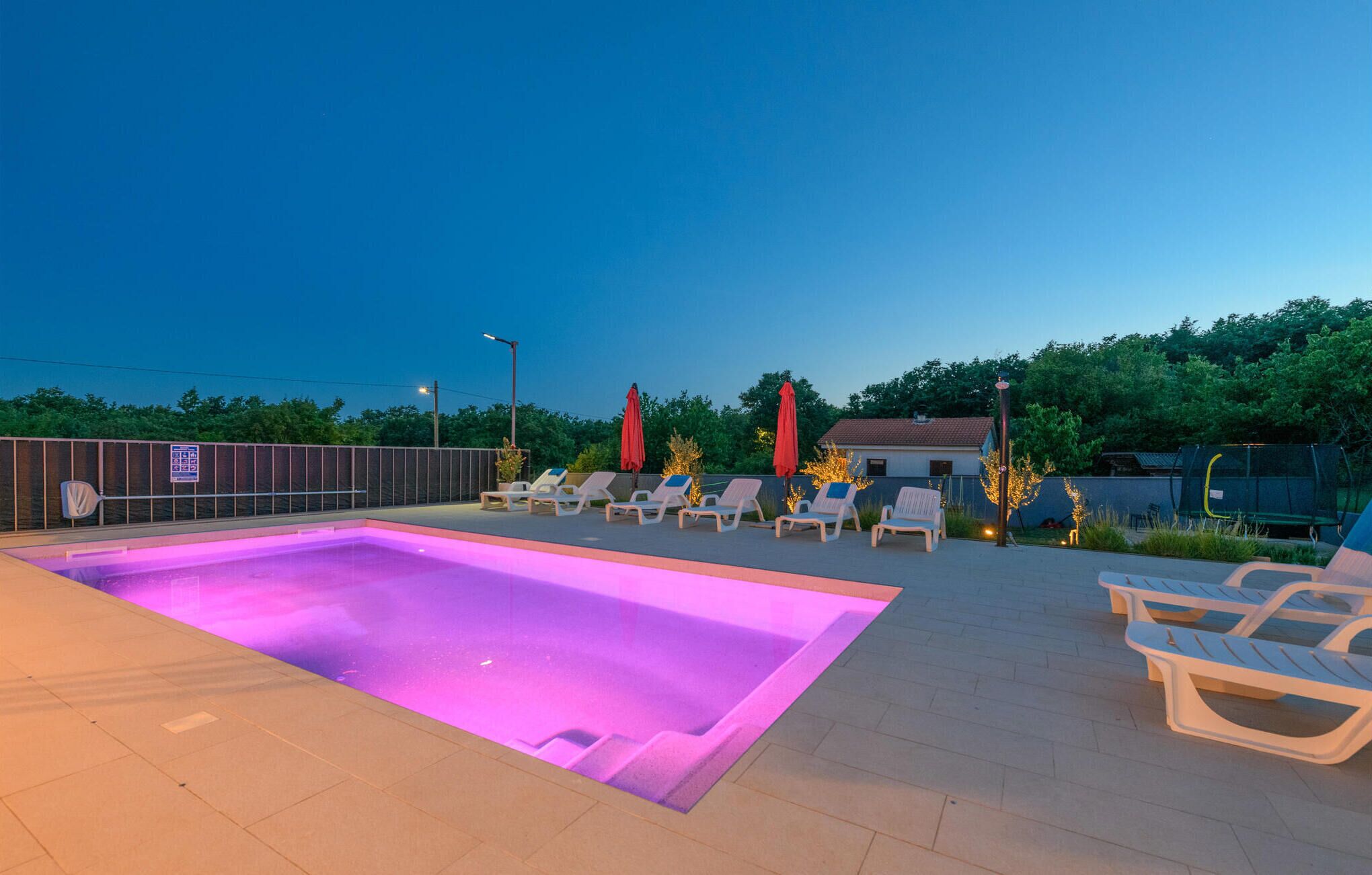 An outdoor swimming pool with lounge chairs and umbrellas, surrounded by a fence and trees.