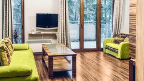 Un salon avec un canapé vert, une table basse en bois et un fauteuil vert agrémenté de coussins à motifs. Un téléviseur à écran plat est fixé au mur, et la pièce est dotée de grandes fenêtres avec des rideaux.