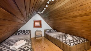 Un dormitorio acogedor en el ático, con paredes y techos inclinados de madera. Hay dos camas individuales con ropa de cama estampada y una pequeña mesita de noche de madera entre ellas.