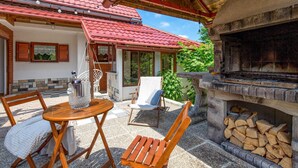 Un área de estar al aire libre con sillas de madera y una mesa, una chimenea de piedra con leña, y un techo de tejas rojas.