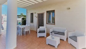 A covered outdoor seating area with white wicker furniture, including a sofa, two armchairs, and a small table. There is a dining table with chairs and a window with curtains.