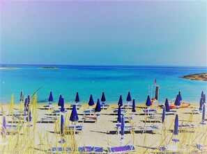 Una playa con hileras de sombrillas azules y sillas de descanso, un mar azul y cristalino, y una isla a lo lejos.