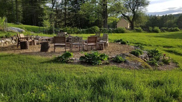 A rustic outdoor seating area with wooden furniture and stone accents, surrounded by lush greenery and a stone wall. - Fabulous, restful, studio with AC in charming Hopkinton (Hopkinton)