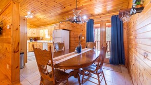 A wooden cabin interior with a dining area featuring a wooden table and chairs, a kitchen with wooden cabinets, and a ceiling with wooden beams.
