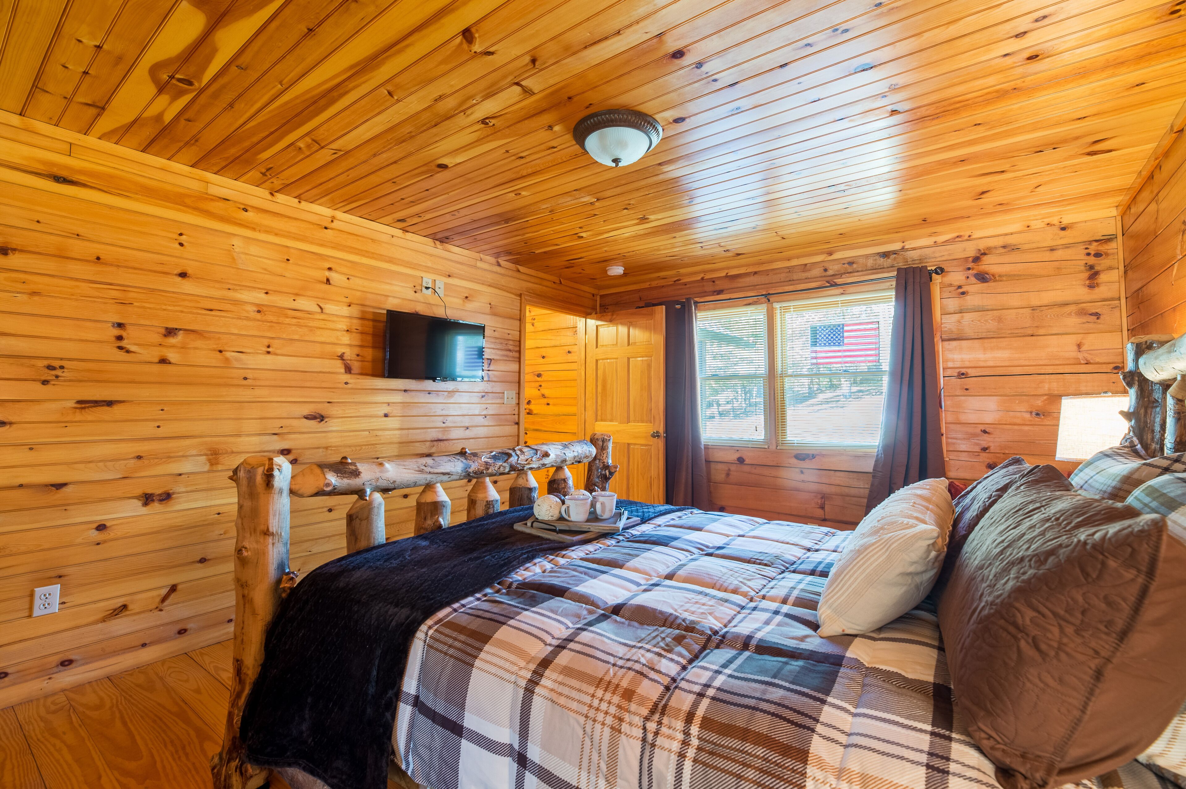 A cozy log cabin bedroom with a plaid bedspread, wooden walls, a mounted television, and a window with curtains.