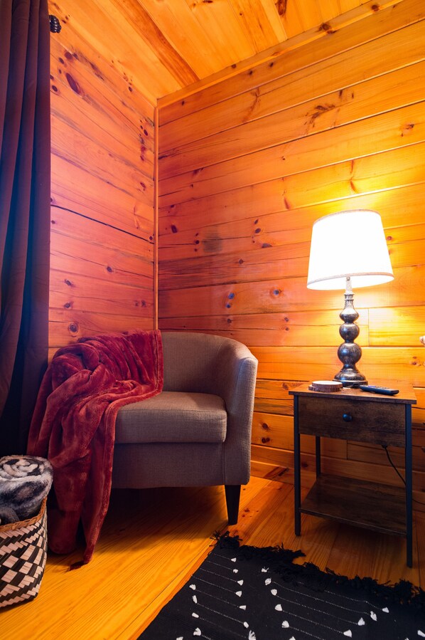 A cozy room with wooden walls, a grey armchair, a red blanket, a lamp on a wooden side table, and a patterned rug. - Relax and Unwind! Secluded Cabin Getaway in the Smokies (Sevierville)