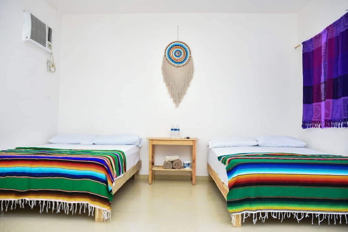 Una habitación con dos camas, cada una con una manta a rayas de colores, una mesita de noche de madera con una botella de agua, y un cuadro colgado en la pared sobre las camas.