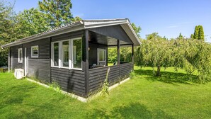 A small, dark wooden cabin with a covered porch, surrounded by lush green grass and trees. - 1 bedroom nice home in Eskebjerg (Eskebjerg)