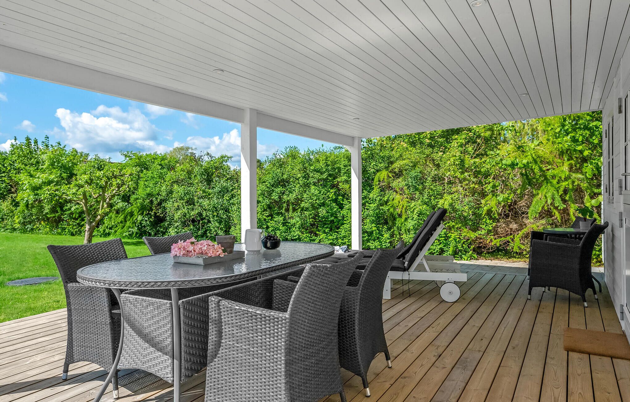A covered patio with a round table and wicker chairs, surrounded by greenery.
