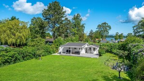 Une maison à deux étages dotée d’un patio couvert et d’une pelouse bien entretenue, entourée d’arbres et offrant une vue sur un lac.