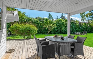 Een overdekt terras met een ronde tafel en rieten stoelen, omgeven door een houten terras en een weelderige tuin.