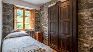 A bedroom with a stone wall, a large wooden wardrobe, a bed with a quilt, and a window with wooden shutters. - Amazing home in Loco di Rovegno (Loco di Rovegno)