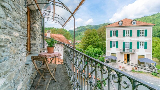 A balcony with a stone wall, a wooden chair, a potted plant, and a view of a two-story building with green shutters. - Amazing home in Loco di Rovegno (Loco di Rovegno)