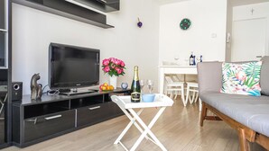 Un salon moderne avec un téléviseur à écran plat, une petite table sur laquelle se trouvent une bouteille et des verres, un canapé agrémenté d’un coussin à motifs floraux, et un coin repas à l’arrière-plan.