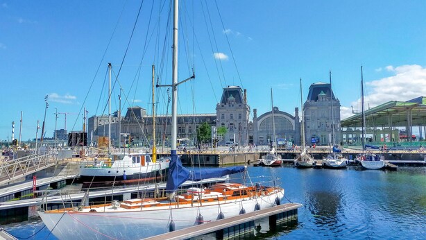 Un port de plaisance où est amarré un grand voilier, entouré d’autres bateaux et avec un bâtiment historique en arrière-plan.