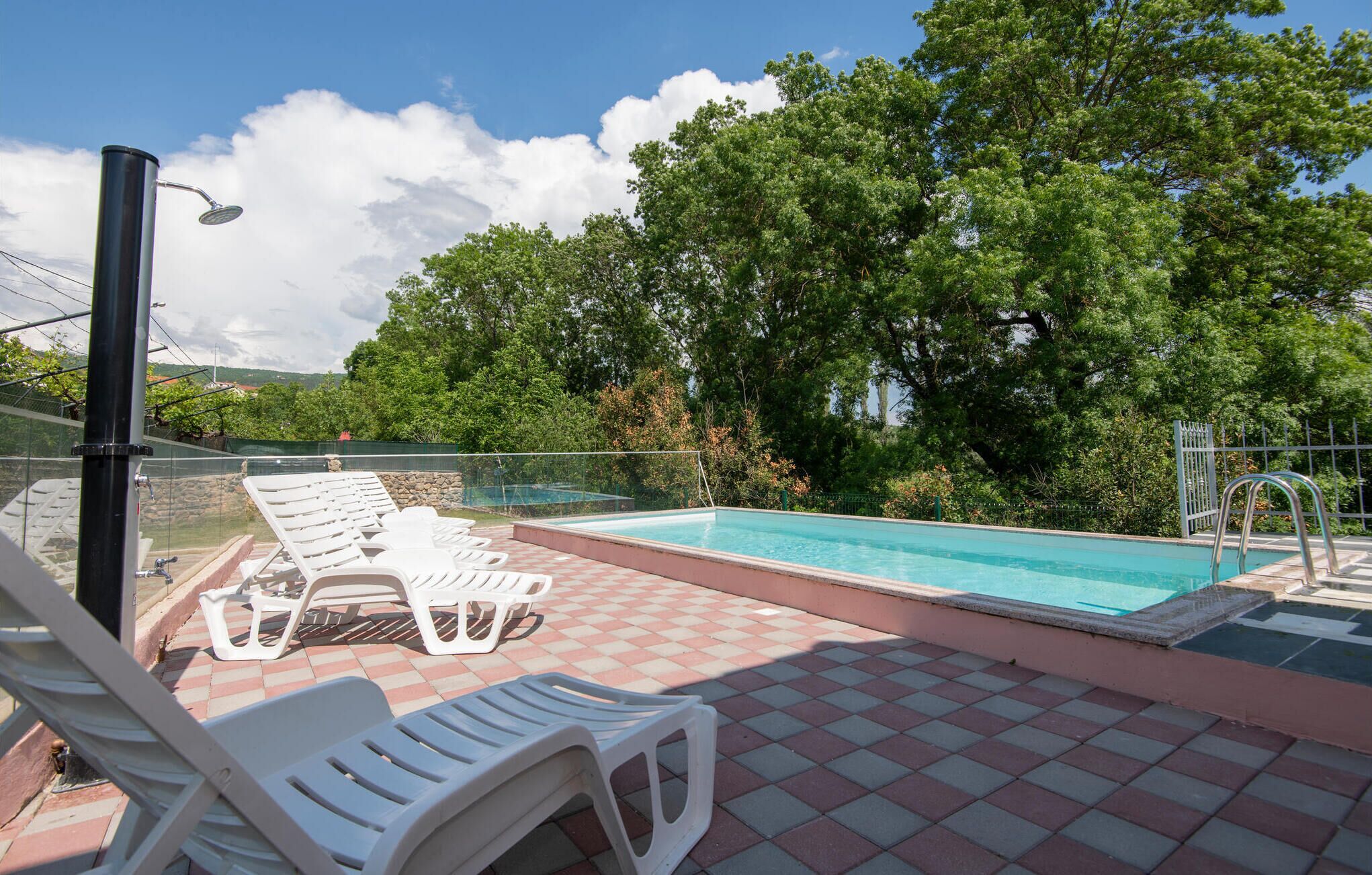 A swimming pool with a tiled deck, surrounded by trees and a clear sky.