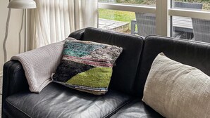 A black leather sofa with a colorful knitted throw pillow and a plain pillow, a white lamp, and a view of a garden through a window. - Pet friendly home in Hvide Sande with sauna (Hvide Sande)