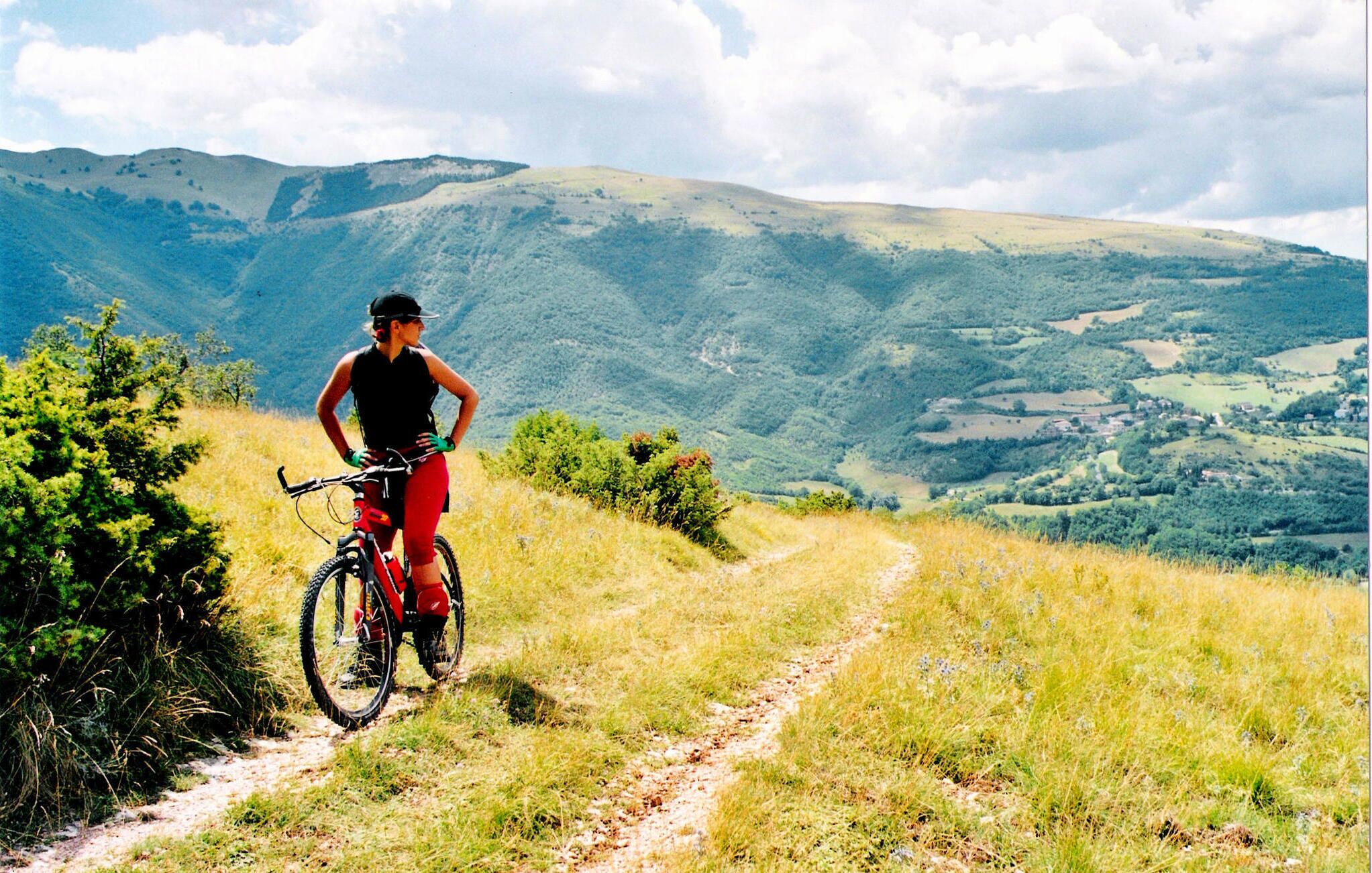 Une personne à vélo tout-terrain sur un sentier de terre, avec une vue panoramique sur des collines et un petit village au loin.