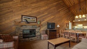 A cozy loft-style living room with a stone fireplace, a television, and a dining area with a kitchen in the background. - Best Location and layout at 7th Mountain 1024 SF 2 Bed/2 Bath - Pet Friendly (Bend)