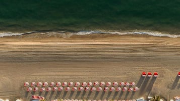 An aerial view of a beach with rows of beach umbrellas and chairs, a sandy shore, and a calm sea.