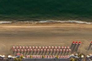 An aerial view of a beach with rows of beach umbrellas and chairs, a sandy shore, and a calm sea.