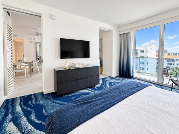 A hotel room with a large bed, a flat-screen TV mounted on the wall, a dark wooden dresser, and a view of the city through large windows.