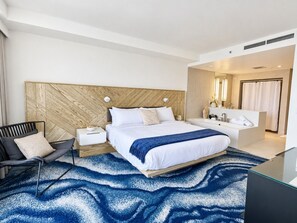 A modern hotel room with a large bed, a sitting area with a chair, a bedside table, and a bathroom with a bathtub and sink.