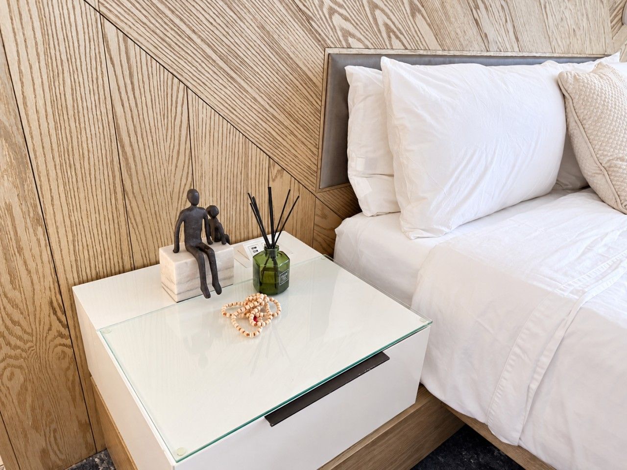 A neatly arranged bedroom with a glass-top nightstand, a bed with white bedding, and a wooden headboard.