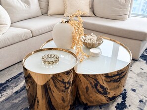 Een woonkamer met een beige bank, twee ronde salontafels met gouden accenten en decoratieve objecten.