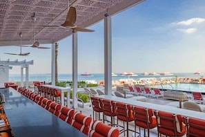 Een zwembadgedeelte met een lange tafel en rode stoelen, met uitzicht op een zwembad en strand met parasols.