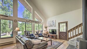 A spacious living room with a leather sofa, a coffee table, and a TV. There are large windows offering a view of trees. A fireplace is visible on the right side. - 'The River Home' Cabin w/ Yard: Near Wolf Creek! (Pagosa Springs)