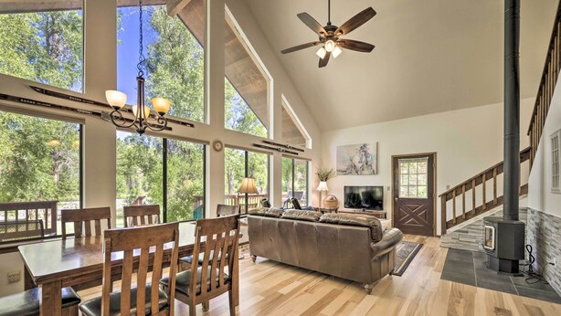 A spacious living room with a high ceiling, large windows, a fireplace, and a dining area with wooden furniture. - 'The River Home' Cabin w/ Yard: Near Wolf Creek! (Pagosa Springs)