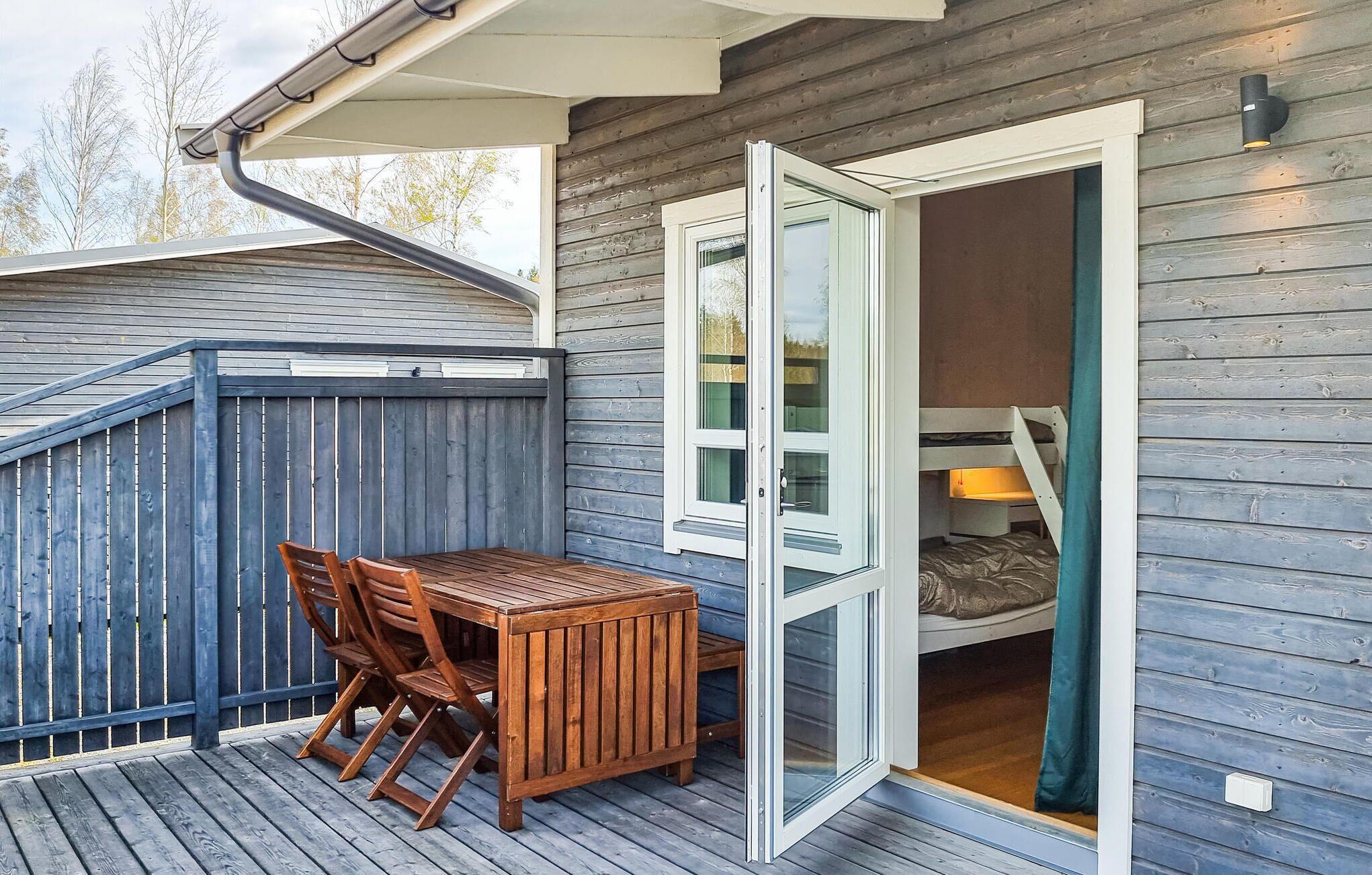 Eine Holzterrasse mit Tisch und Stühlen, eine Glasschiebetür zu einem Raum mit einem Etagenbett und ein blaues Geländer.