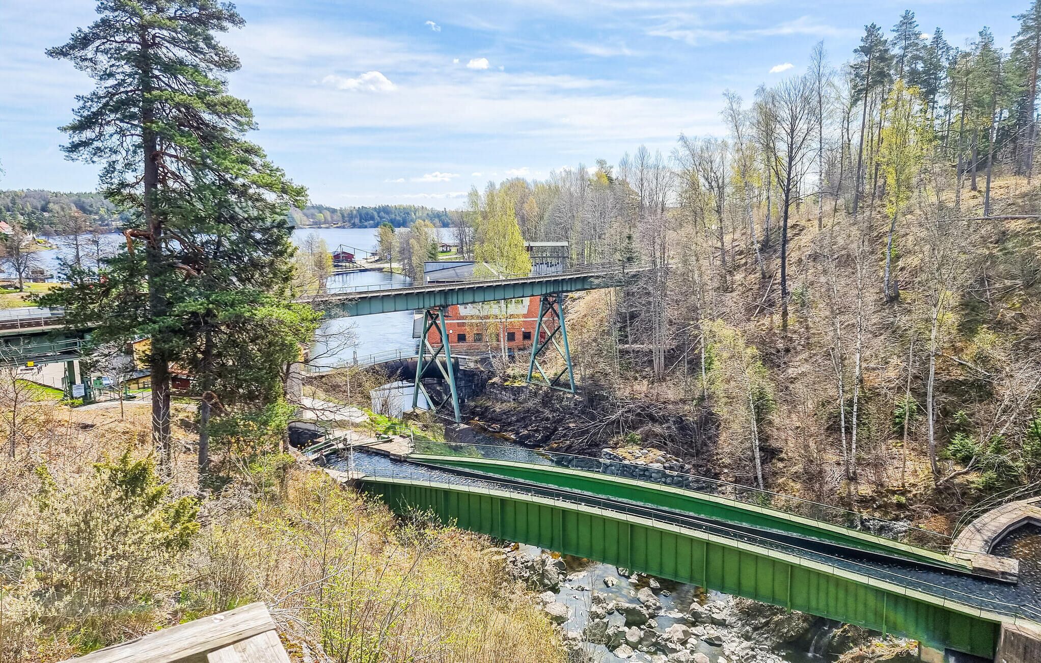 A green pedestrian bridge over a river with a small waterfall, surrounded by trees and a distant building.