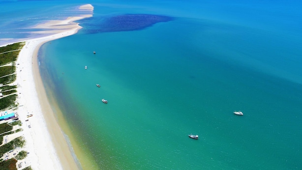 Une vue aérienne d’une plage aux eaux turquoise et limpides, avec plusieurs bateaux et une rive sablonneuse parsemée de quelques constructions.