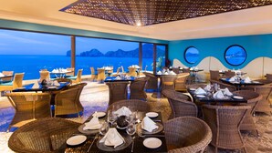 A restaurant with wicker chairs and tables set for dining, overlooking a scenic view of the sea and mountains.