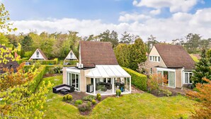Une maison moderne dotée d’une véranda en verre, d’un jardin bien entretenu et située à proximité d’une maison traditionnelle.
