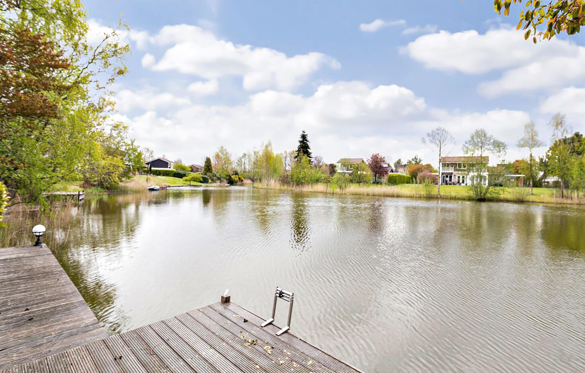 Een rustig meer met een houten steiger die in het water reikt, omringd door bomen en huizen op de achtergrond.