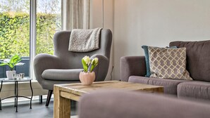 Un salon moderne avec un fauteuil gris, une table basse en bois et un canapé agrémenté de coussins à motifs. Une plante en pot et un vase de fleurs sont posés sur la table basse.