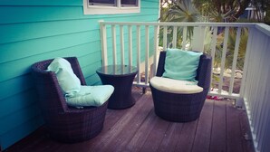 Een veranda met twee rieten stoelen en een kleine tafel, een witte balustrade, en een blauw huis op de achtergrond.