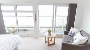 Quarto com cama, sofá, uma mesinha com uma planta e grandes janelas com vista para o mar.