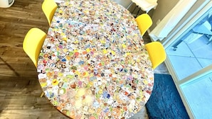Een ronde tafel met een kleurrijk, cartoonesk patroon, omringd door gele stoelen, in een kamer met een houten vloer en een groot raam.
