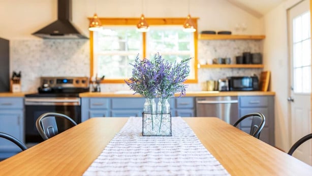 A kitchen with a wooden table, a vase with flowers, and a window with natural light.