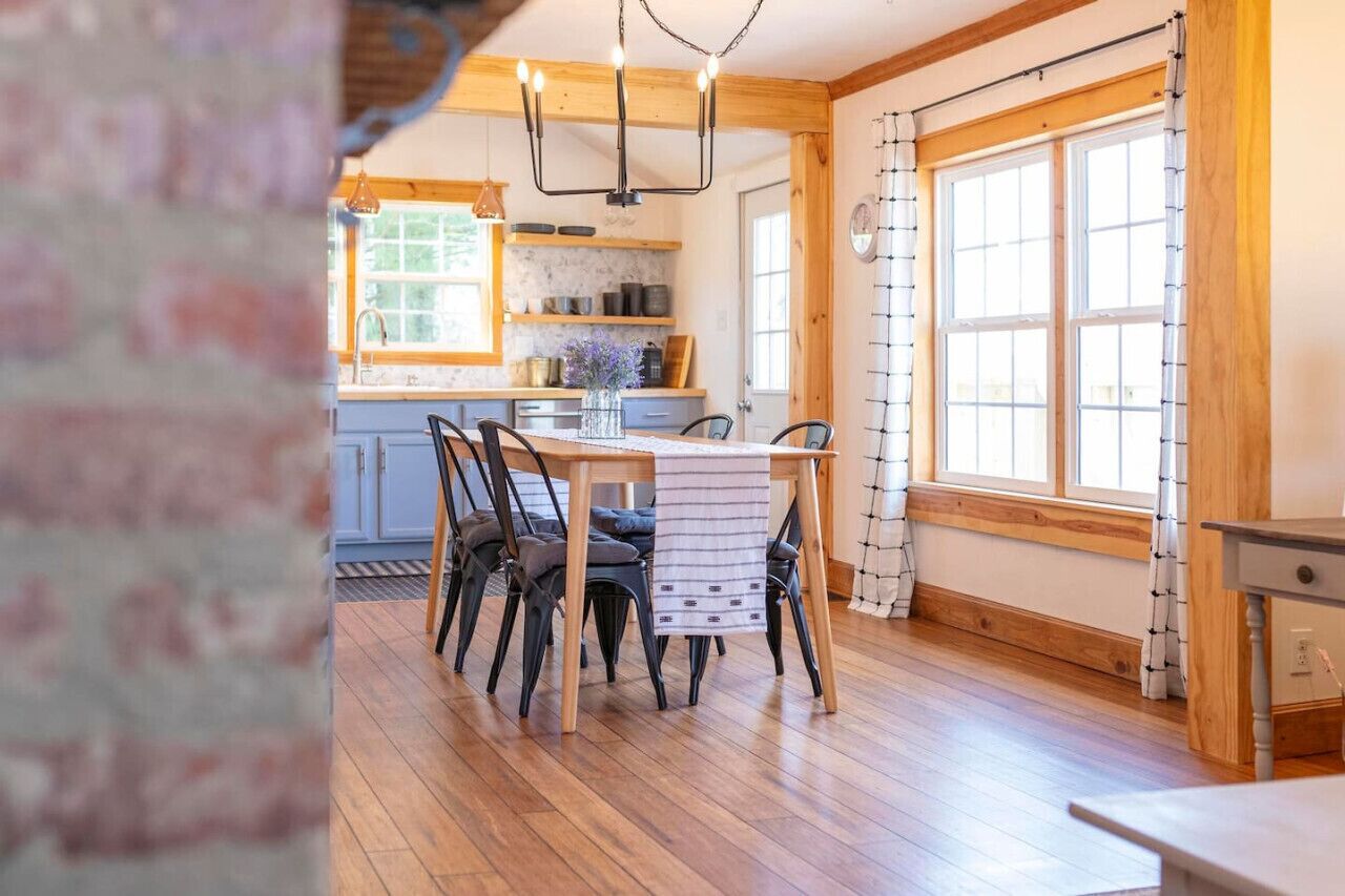 Une table à manger en bois, entourée de chaises, un lustre au plafond et, au fond, une cuisine.