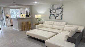 A modern living room with a sectional sofa, a dining area with a kitchen in the background, and a large abstract painting on the wall. - Beautiful home on a deep water canal leading to the Gulf of America. (New Port Richey)