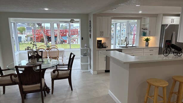 A modern kitchen with white cabinetry, a central island with stools, and large windows offering a view of outdoor play equipment. - Beautiful home on a deep water canal leading to the Gulf of America. (New Port Richey)