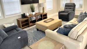 Ein Wohnzimmer mit einem beigen Sofa, einem grauen Sessel, einem Couchtisch aus Holz und einem an der Wand montierten Fernseher.