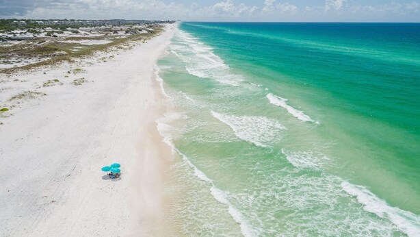 Eine Vogelperspektive zeigt einen Strand mit blauem Himmel, sandigem Ufer und sanften Wellen.